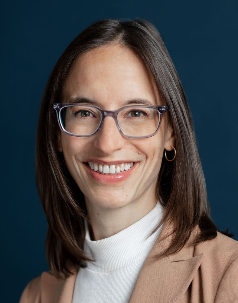 Woman with straight brown hair, wearing glasses, a beige blazer, and a white turtleneck, smiling in front of a dark blue background.